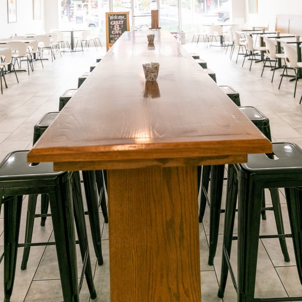 Long wooden communal table with black stools in a bright, empty restaurant dining room.