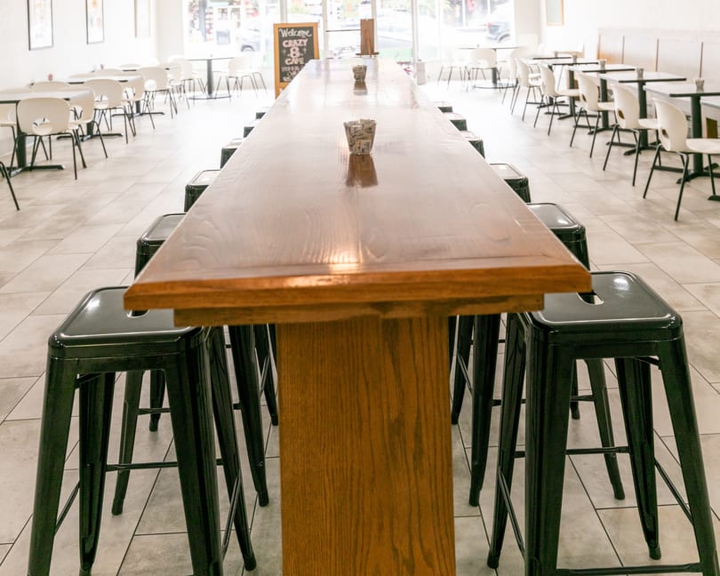 Long wooden communal table with black stools in a bright, empty restaurant dining room.