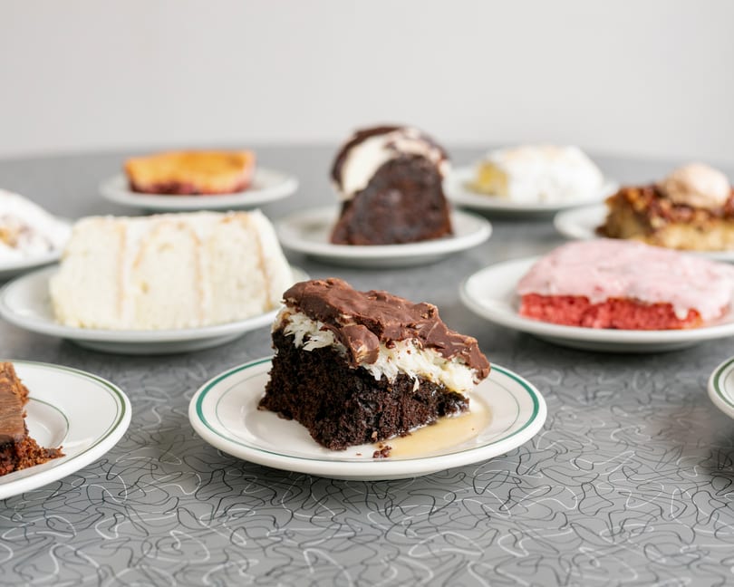 Assortment of cake slices on white plates with green rims on a gray patterned table.