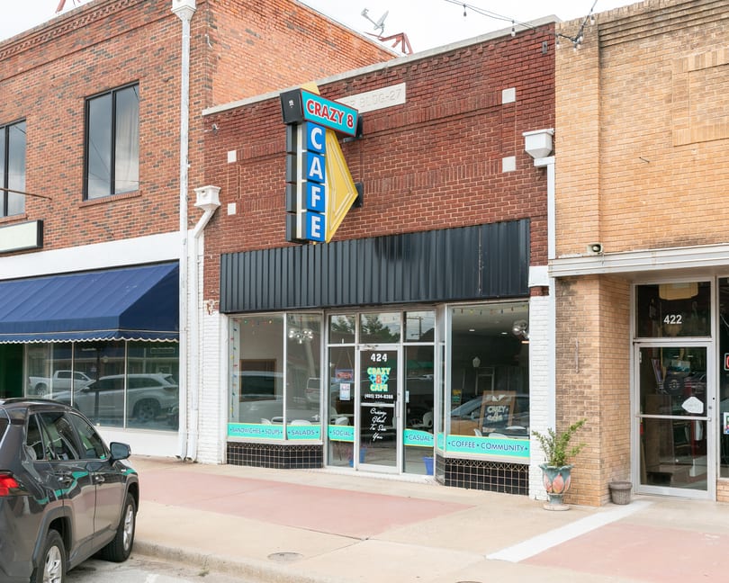 Exterior of Crazy 8 Cafe, a brick building with a blue and yellow sign, storefront windows, and a parked car.