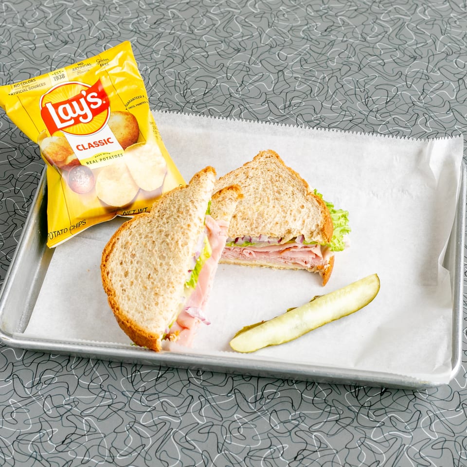 A ham sandwich with lettuce and red onion, a pickle spear, and Lay's chips on a tray.