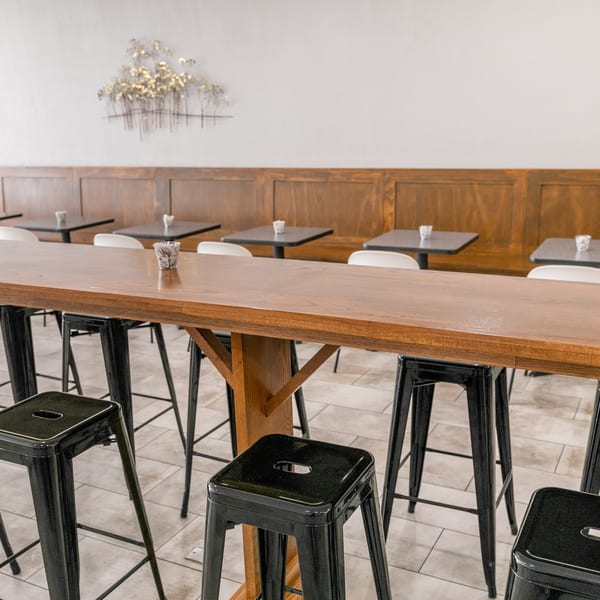 Long wooden communal table with black stools, and smaller tables with white chairs against a wooden wall.