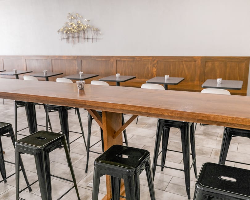 Long wooden communal table with black stools, and smaller tables with white chairs against a wooden wall.