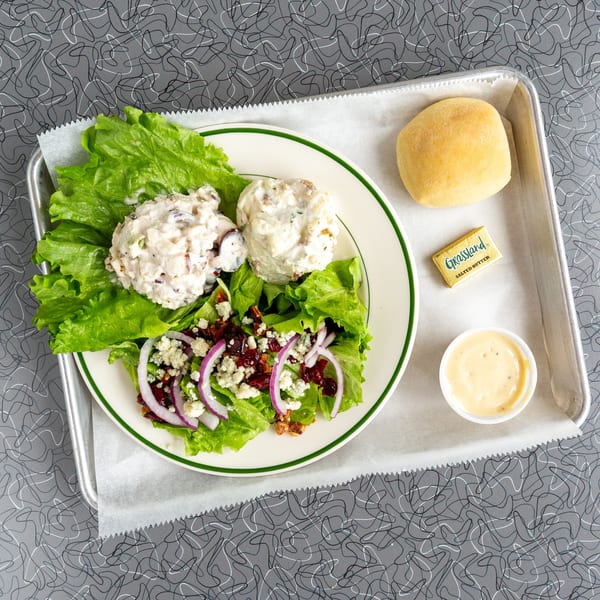 Chicken salad plate with a side salad, roll, butter, and dressing on a metal tray.