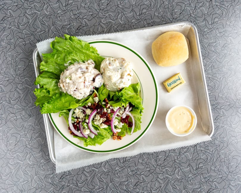 Chicken salad plate with a side salad, roll, butter, and dressing on a metal tray.
