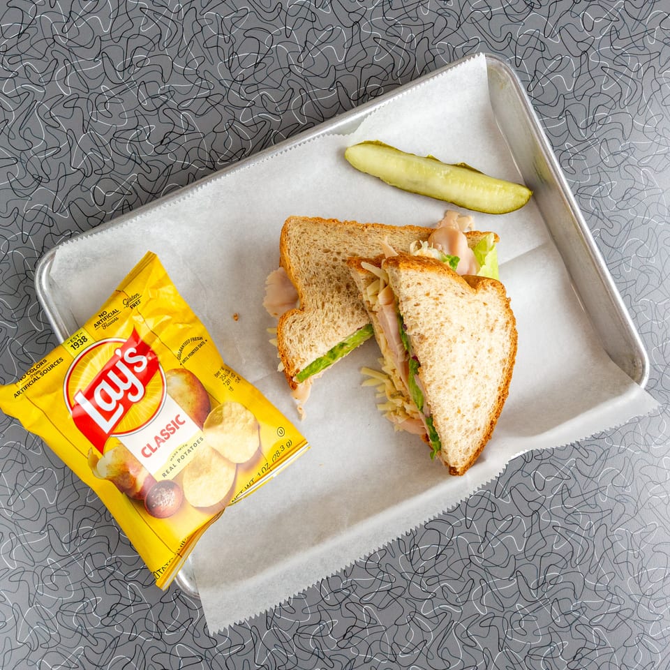 Turkey sandwich with lettuce, cheese, a pickle spear, and a bag of Lay's chips on a tray.