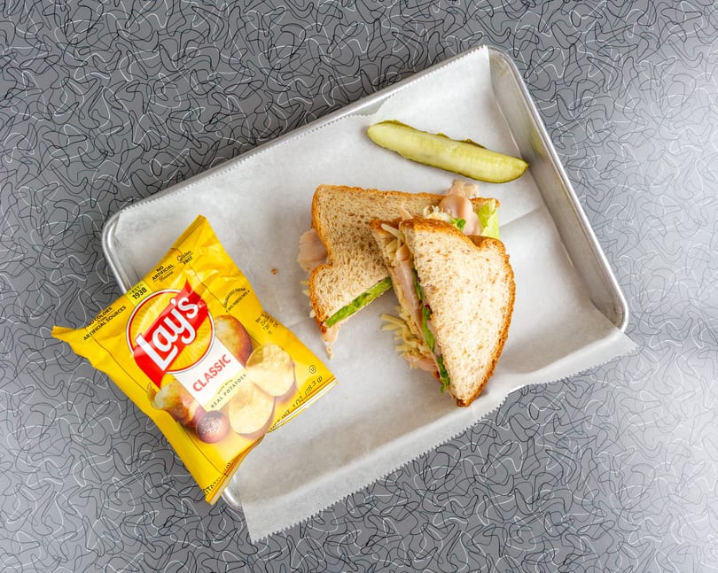 Turkey sandwich with lettuce, cheese, a pickle spear, and a bag of Lay's chips on a tray.