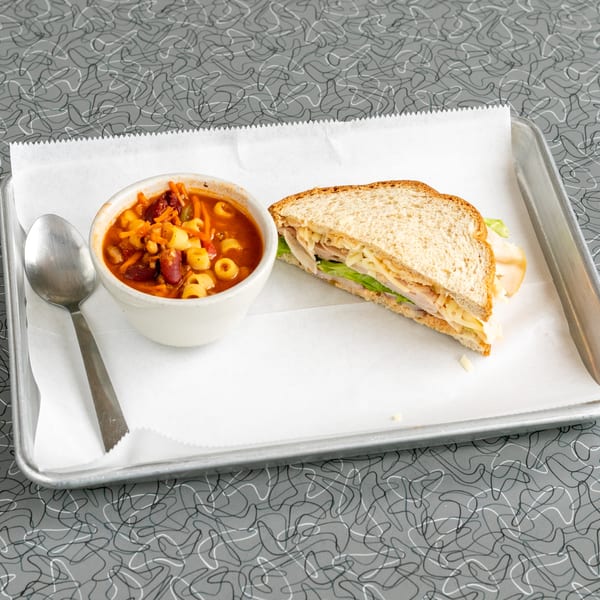 A bowl of minestrone soup with a spoon and a turkey sandwich on a metal tray.