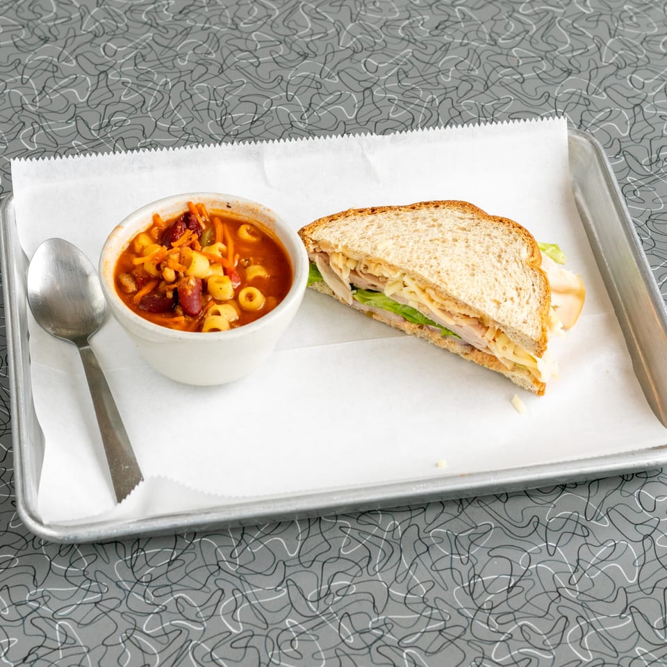 A bowl of minestrone soup with a spoon and a turkey sandwich on a metal tray.