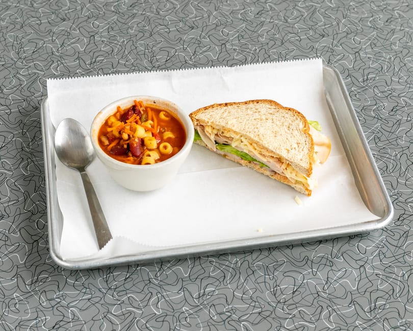 A bowl of minestrone soup with a spoon and a turkey sandwich on a metal tray.