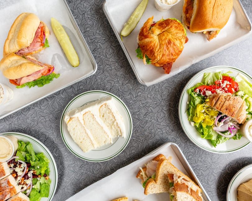 Overhead view of various sandwiches, salads, and a slice of cake on a patterned gray table.