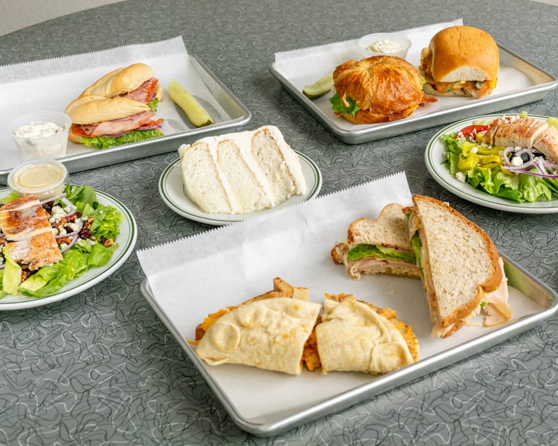 A full table of sandwiches, salads, and a slice of cake on a retro patterned table.