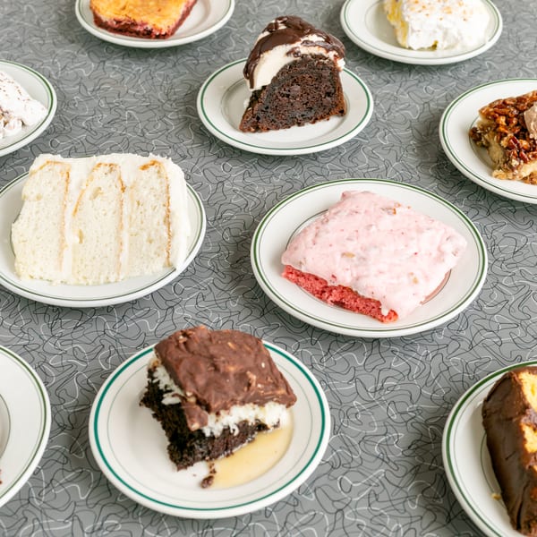 Nine slices of various cakes and desserts on white plates with green rims, on a gray patterned table.