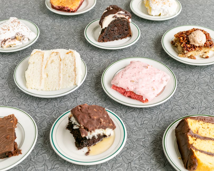Nine slices of various cakes and desserts on white plates with green rims, on a gray patterned table.