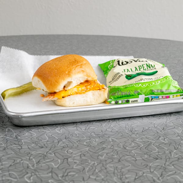 A chicken slider with melted cheese, a pickle spear, and a bag of Miss Vickie's Jalapeño chips.