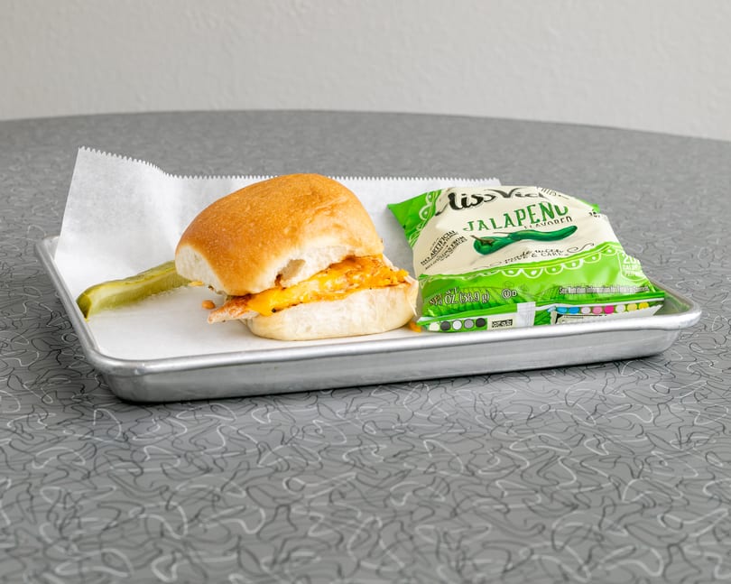 A chicken slider with melted cheese, a pickle spear, and a bag of Miss Vickie's Jalapeño chips.