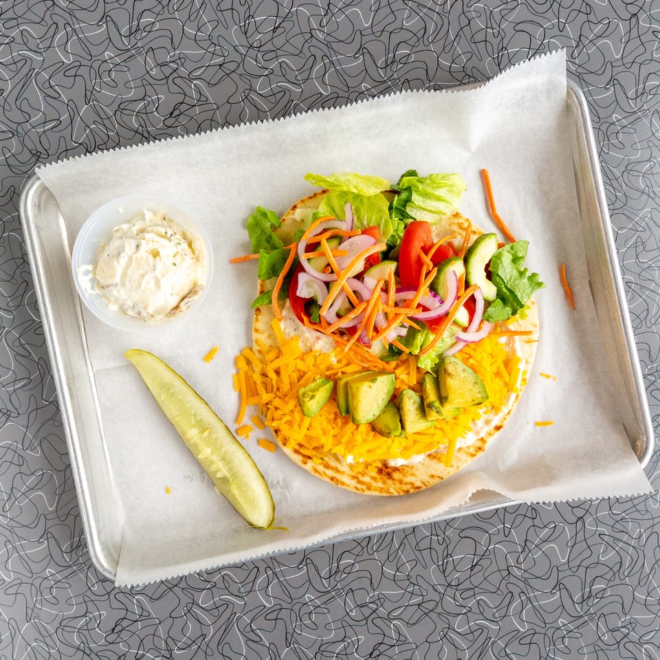 A pita bread with lettuce, tomatoes, onions, carrots, avocado, and shredded cheese, with a side of potato salad and a pickle spear.