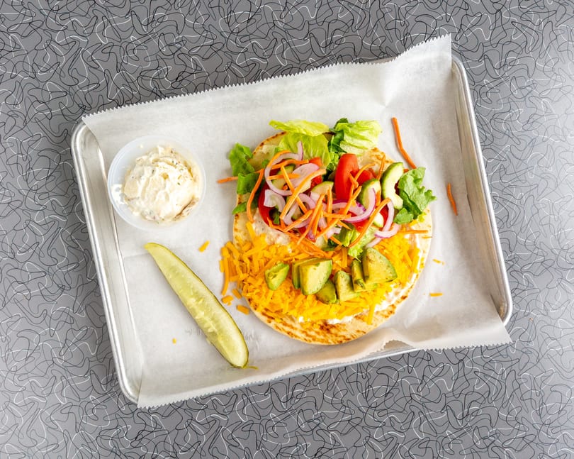 A pita bread with lettuce, tomatoes, onions, carrots, avocado, and shredded cheese, with a side of potato salad and a pickle spear.