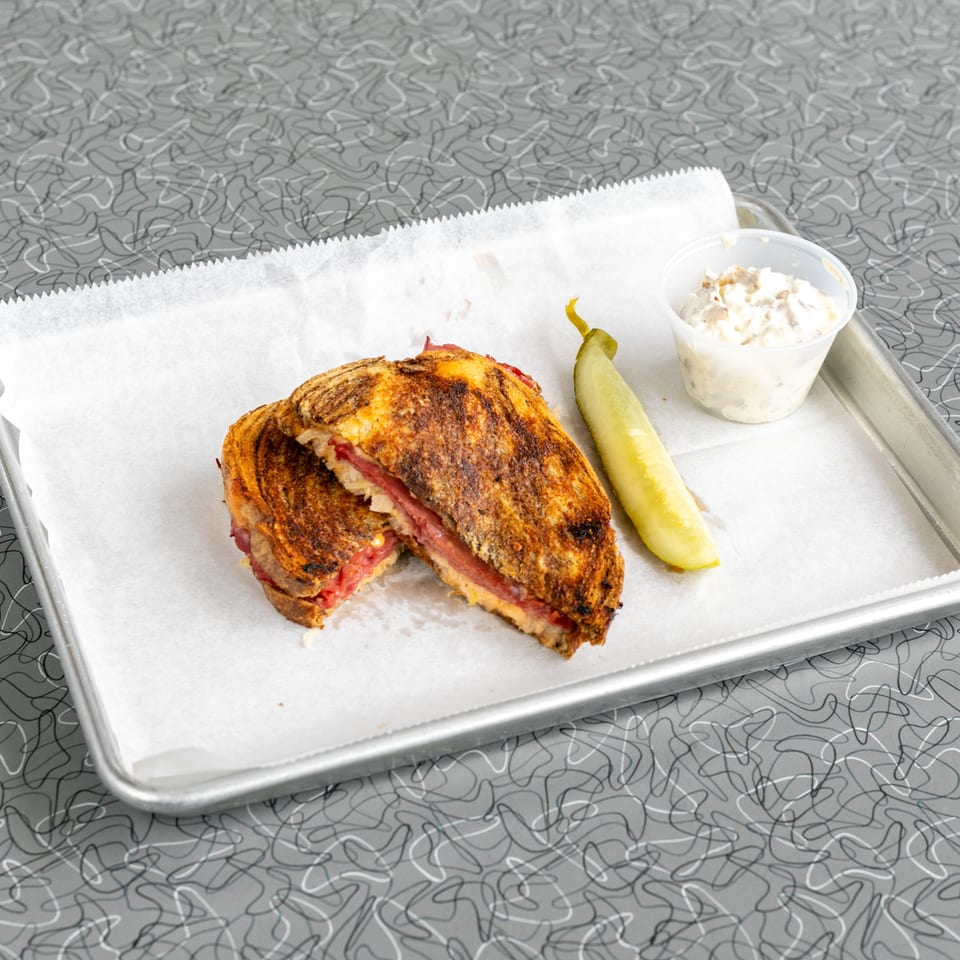 A grilled sandwich, cut in half, with a pickle spear and coleslaw on a metal tray.