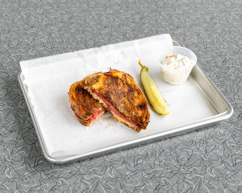 A grilled sandwich, cut in half, with a pickle spear and coleslaw on a metal tray.