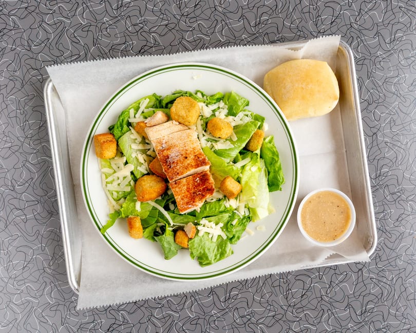 Chicken Caesar salad with croutons, shredded cheese, a dinner roll, and dressing on a tray.