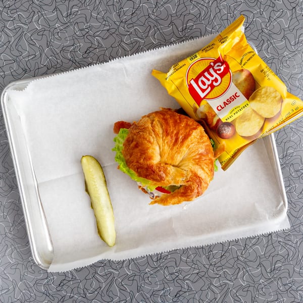 Croissant sandwich with lettuce, tomato, and bacon, a pickle spear, and Lay's chips on a tray.