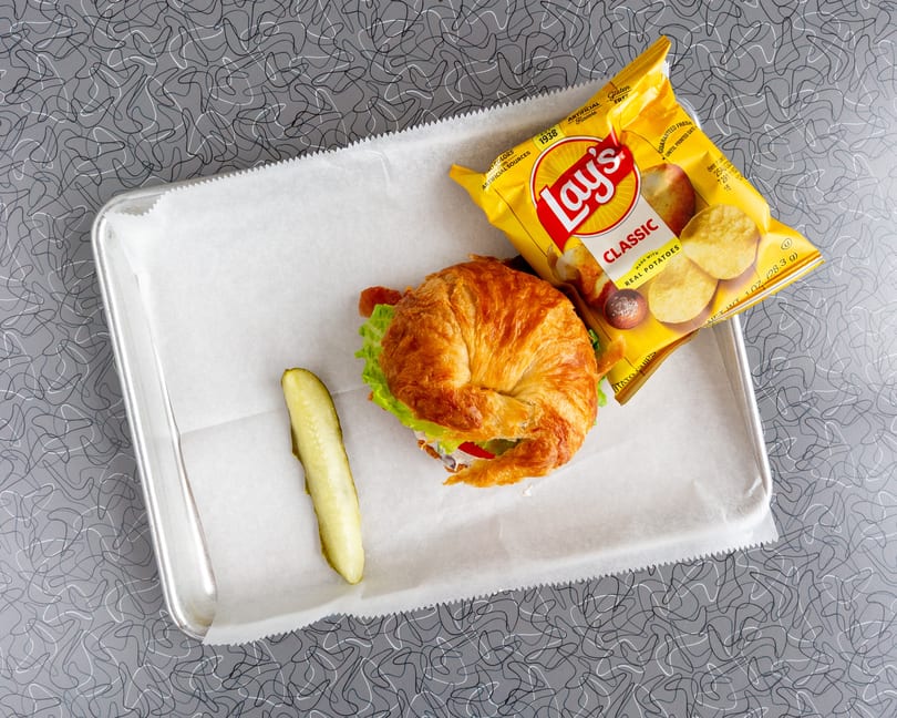 Croissant sandwich with lettuce, tomato, and bacon, a pickle spear, and Lay's chips on a tray.