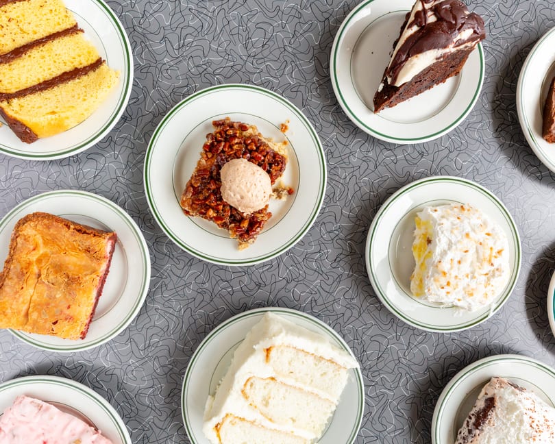 Assortment of dessert slices, including cakes, pies, and brownies, on a diner-style tabletop.
