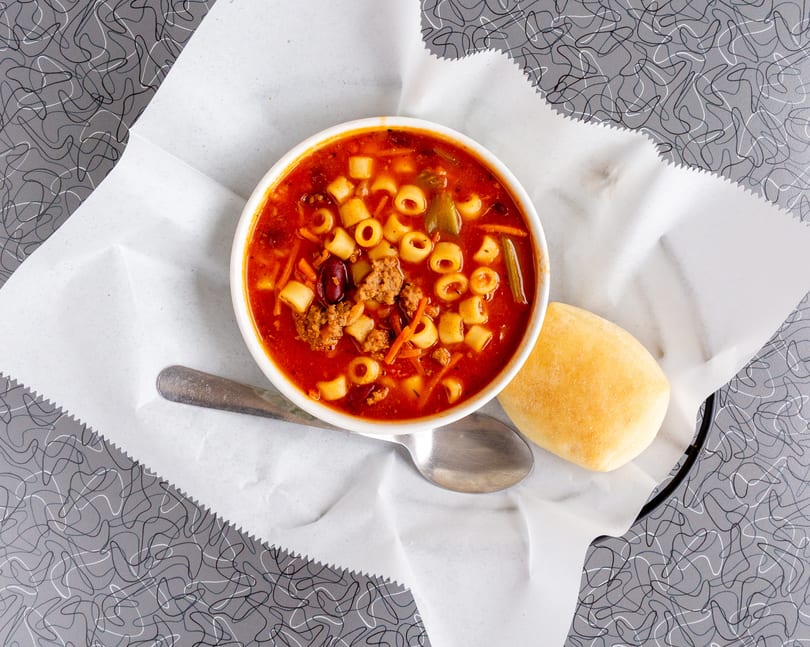 Hearty minestrone soup with pasta, beans, and vegetables, served with a bread roll.