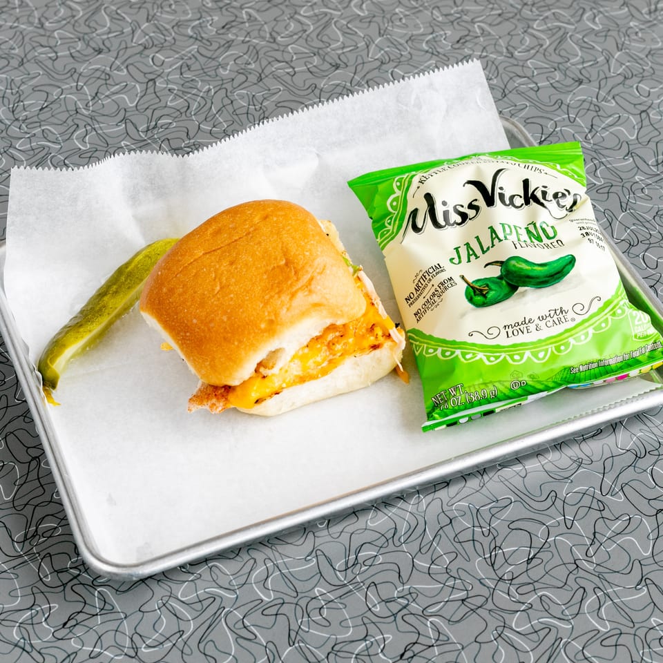 A chicken sandwich with melted cheese, a pickle spear, and a bag of Miss Vickie's Jalapeño chips.
