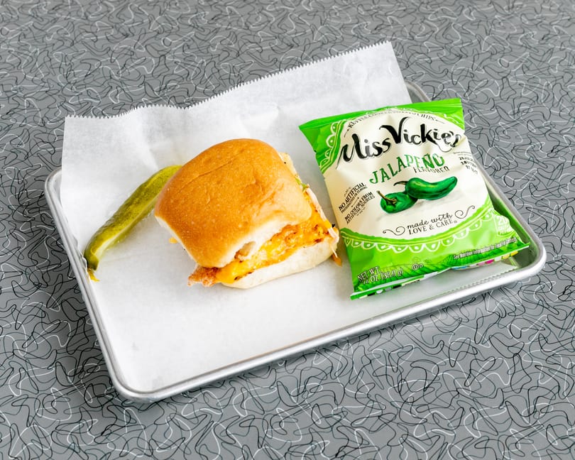 A chicken sandwich with melted cheese, a pickle spear, and a bag of Miss Vickie's Jalapeño chips.