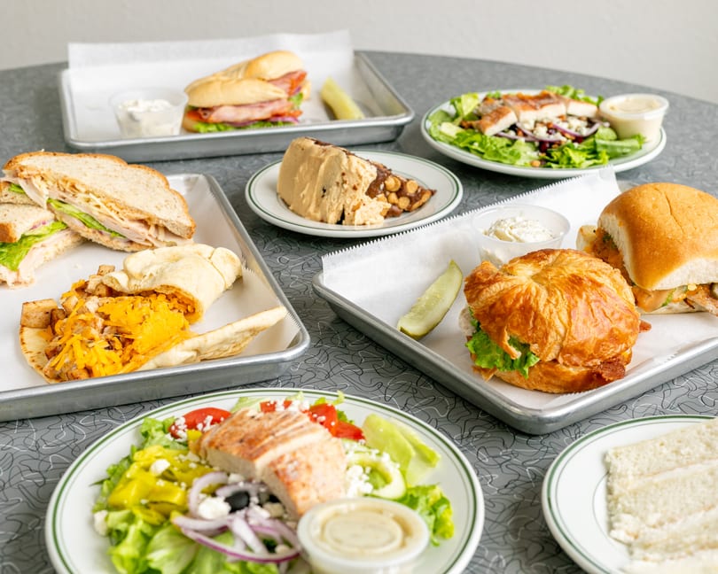 A table filled with various sandwiches, salads, and a dessert on plates and metal trays.
