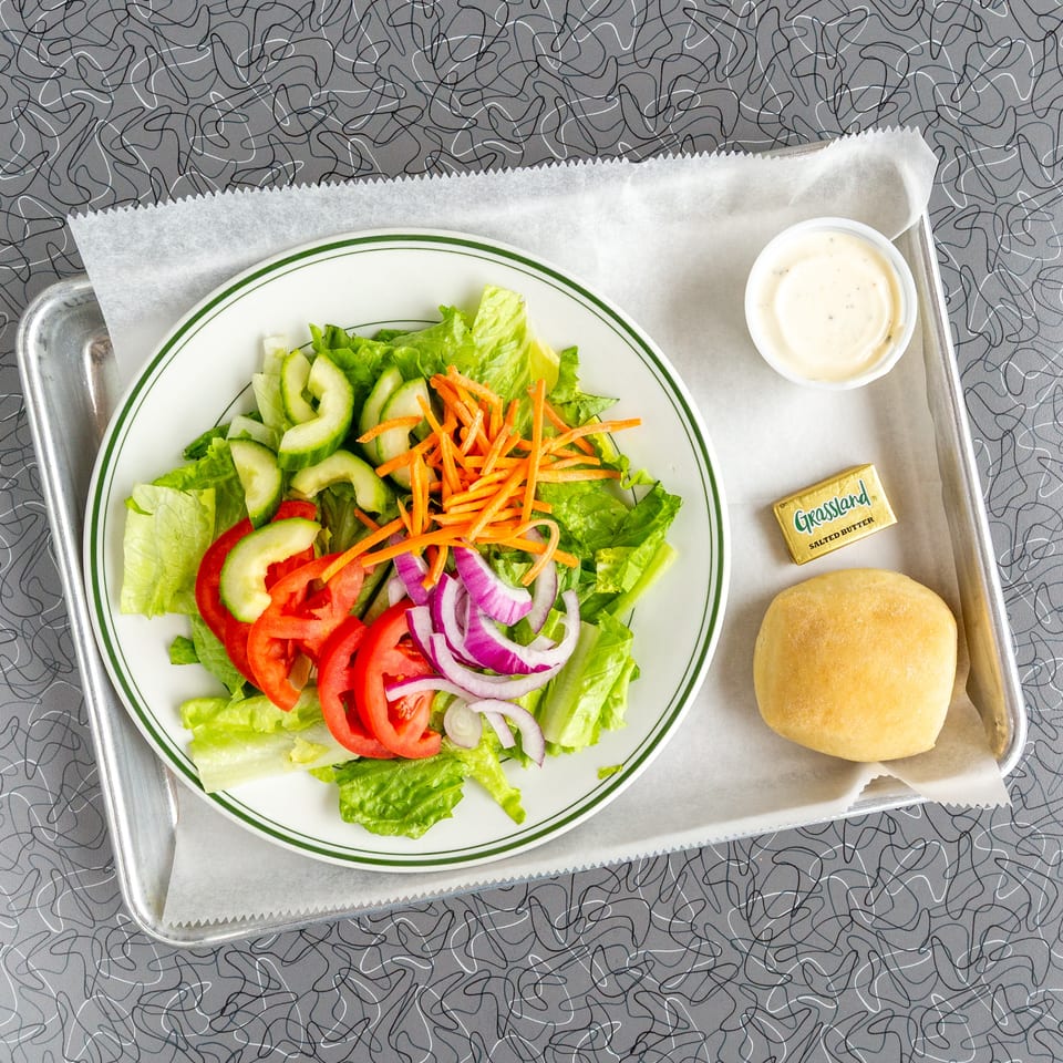 A fresh side salad with lettuce, tomatoes, cucumbers, onions, and carrots, served with a roll and butter.