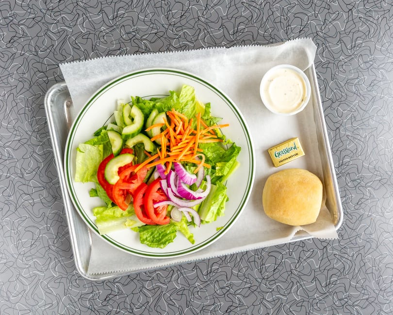 A fresh side salad with lettuce, tomatoes, cucumbers, onions, and carrots, served with a roll and butter.