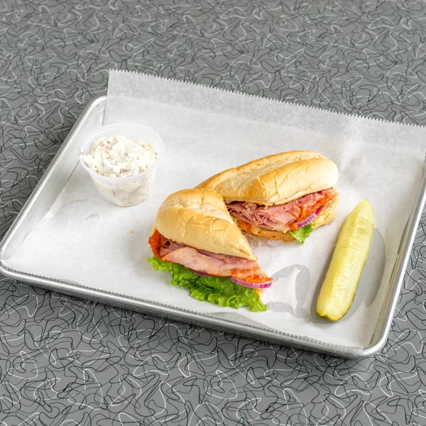 A deli sub sandwich, cut in half, with a side of coleslaw and a pickle spear on a tray.