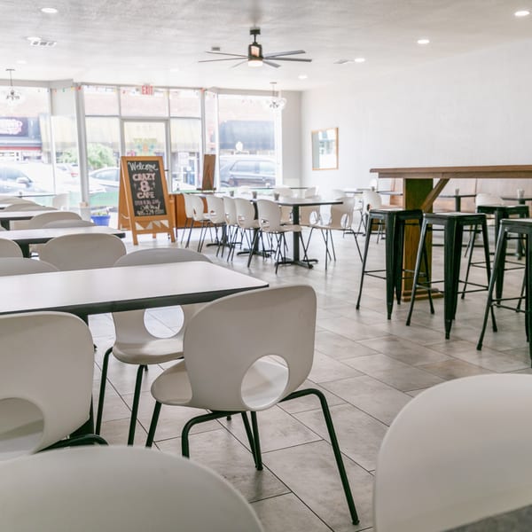 Bright, empty restaurant interior with white tables, chairs, and a long wooden bar with black stools.