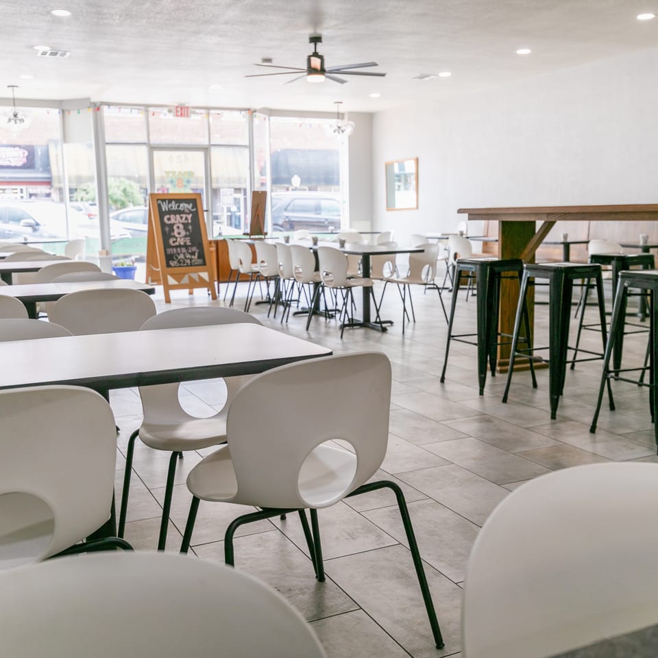 Bright, empty restaurant interior with white tables, chairs, and a long wooden bar with black stools.