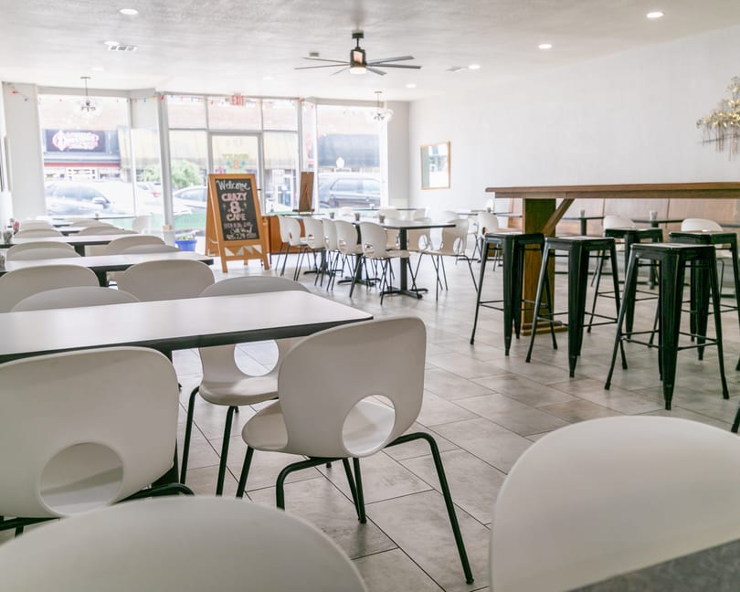 Bright, empty restaurant interior with white tables, chairs, and a long wooden bar with black stools.