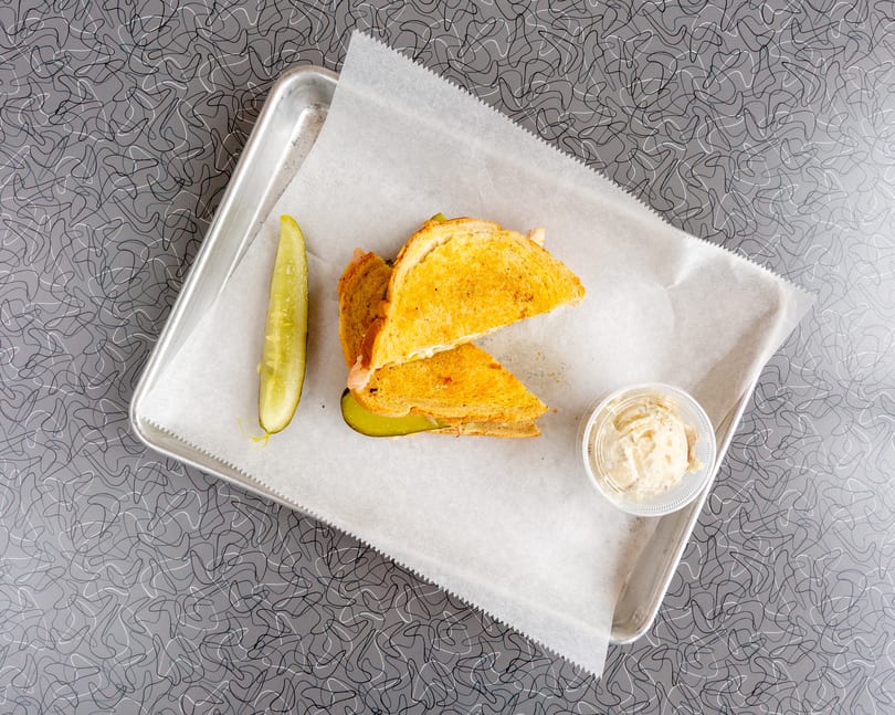 Grilled sandwich halves, a pickle spear, and a side of potato salad on a metal tray.