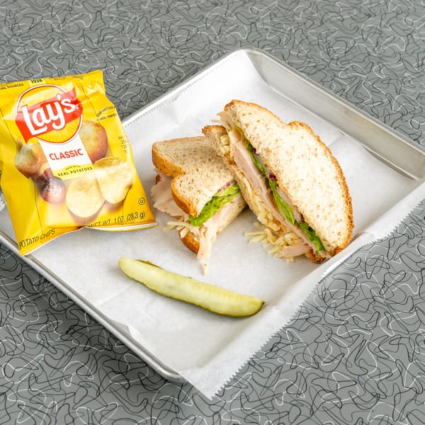 Turkey sandwich with lettuce, cheese, and red onion, served with Lay's chips and a pickle spear.