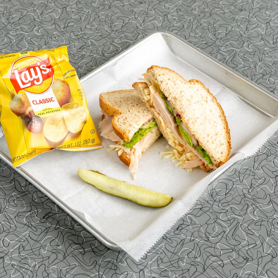 Turkey sandwich with lettuce, cheese, and red onion, served with Lay's chips and a pickle spear.