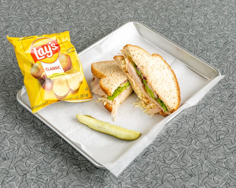 Turkey sandwich with lettuce, cheese, and red onion, served with Lay's chips and a pickle spear.
